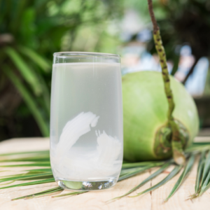 Kokoswater in een glas op het strand. Achter het glas ligt een jonge groene kokosnoot.