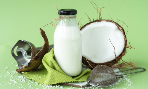Glazen flesje kokosmelk met een halve kokosnoot ernaast, groen achtergrond Hoe maak je Romige natuurlijke Kokosmelk
